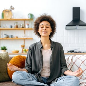 Vertical photo of calm peaceful relaxed african american woman in casual clothes, sitting alone at home in living room on the couch, resting and meditating in the lotus position, smiling